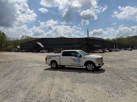 White pickup truck parked on gravel lot in front of large dark hangar building under blue sky with clouds