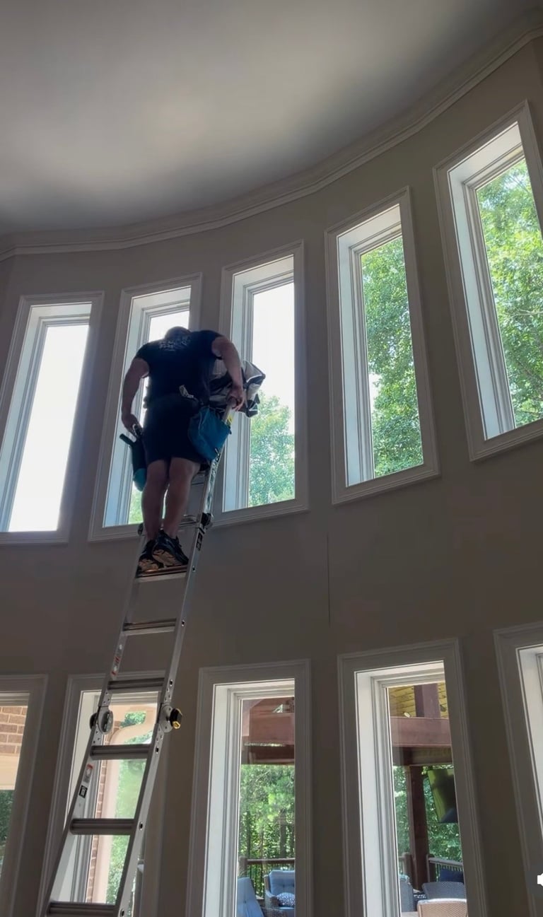 Person on ladder cleaning or maintaining tall windows in a sunlit room with multiple paned windows