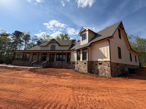Modern farmhouse under construction with tan and brown siding, stone foundation, and red clay driveway on sunny day