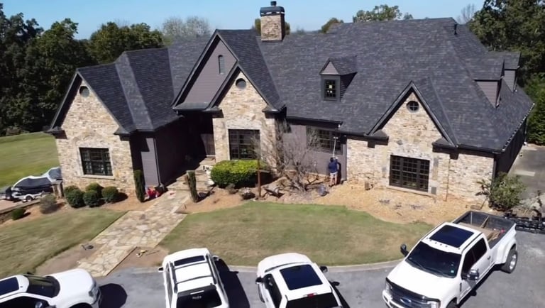 Aerial view of a large two-story stone and dark-roofed house with manicured lawn and white vehicles parked in the driveway