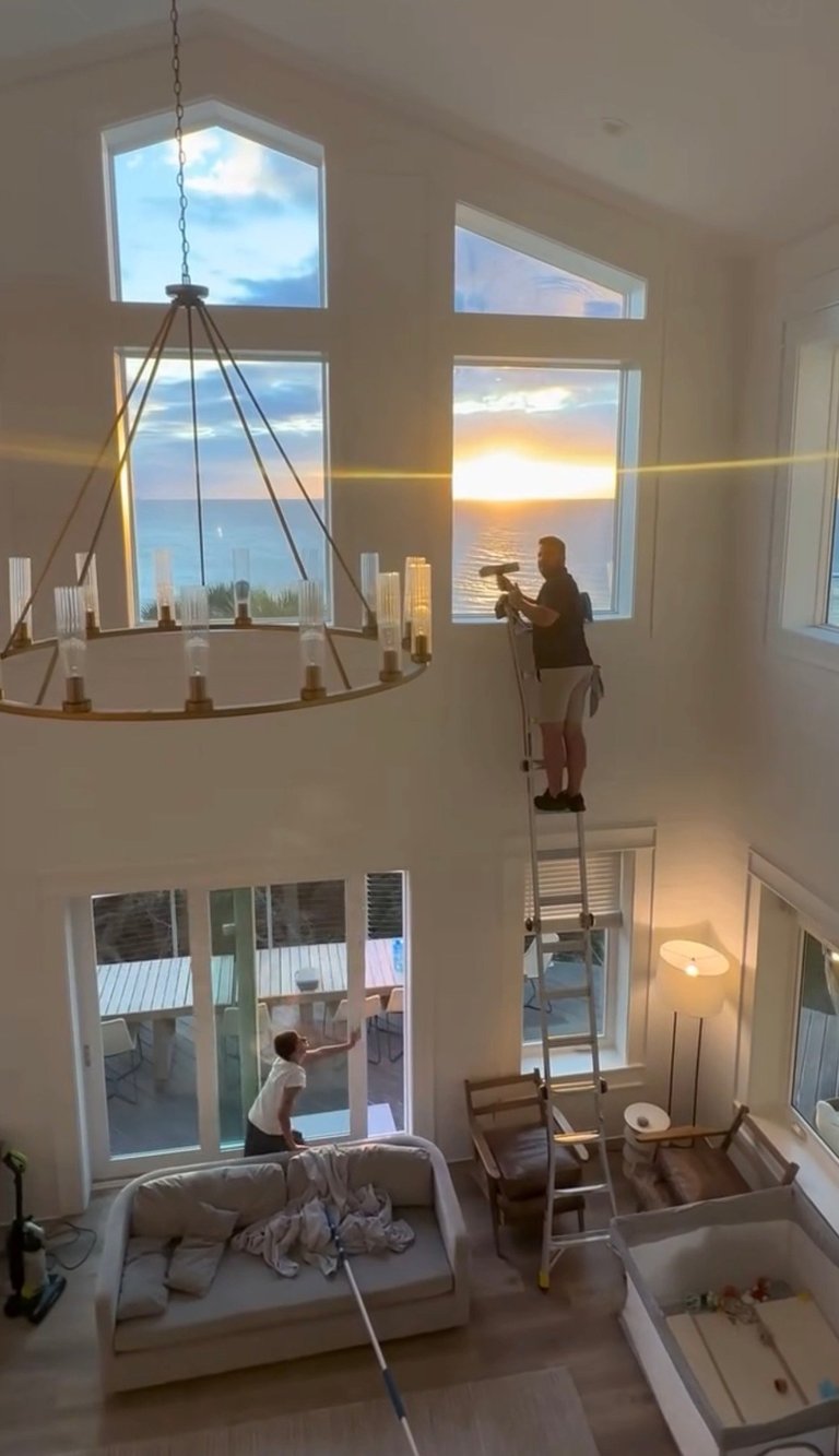 Two people on ladders installing a chandelier in a two-story modern home with large windows overlooking a city and sunset